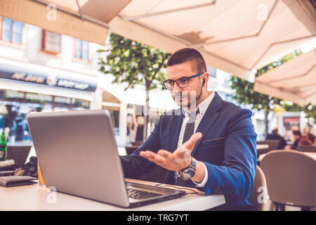 Giovane uomo seduto al caffè strada avente una conversazione su un laptop che indossa una cuffia sulla testa parlando guarda arrabbiato solleva una mano. Foto Stock