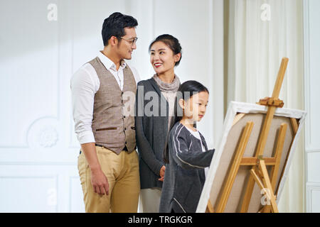 Bella ragazza asiatica con lunghi capelli neri facendo un dipinto su tela mentre i genitori di stare dietro a guardare, focus sui genitori. Foto Stock