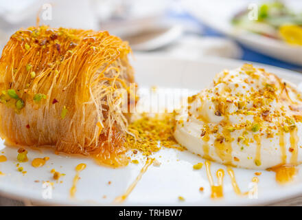 Close up di un greco succulenti dessert. Kataifi greco con miele yogurt e pistacchi grattugiato su una piastra bianca e uno sfondo sfocato. Vie orizzontale Foto Stock