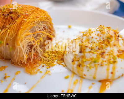 Close up di un greco succulenti dessert. Kataifi greco con miele yogurt e pistacchi grattugiato su una piastra bianca e uno sfondo sfocato. Vie orizzontale Foto Stock