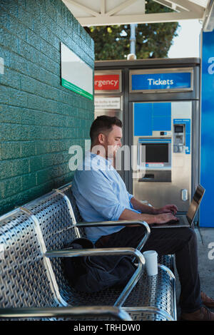 Una vista laterale colpo di una metà degli adulti imprenditore caucasico lavorando sul suo computer portatile mentre si è seduti in attesa del treno alla stazione ferroviaria e piattaforma. Foto Stock