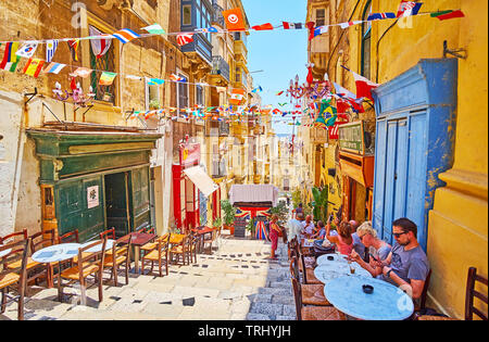 La Valletta, Malta - 19 giugno 2018: Le tabelle di accoglienti bar esterno tratto lungo la discesa di St Lucia street, decorata con colorate ghirlande di bandiera, o Foto Stock