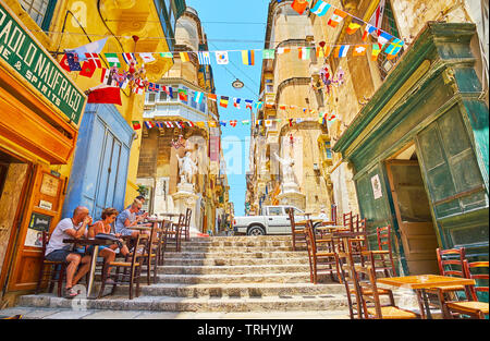 La Valletta, Malta - 19 giugno 2018: la stretta discesa di St Lucia street è rivestito con tabelle di locale bar e caffetterie, decorato con ghirlande di bandiera, su Foto Stock