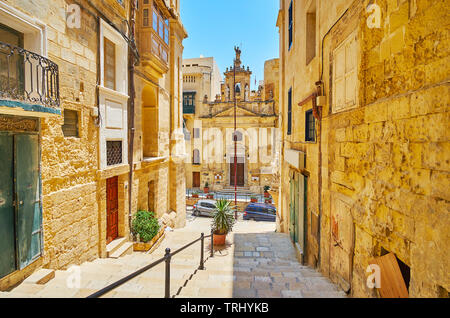 La vista sulla facciata scolpita del borgo medievale di St Lucia chiesa dalla stretta discesa di St Lucia Street, Valletta, Malta. Foto Stock