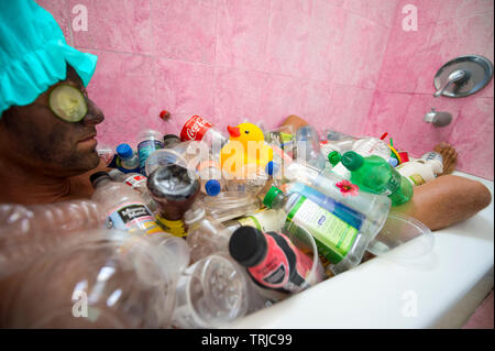 MIAMI - Luglio 2018: un uomo cerca di rilassarsi con una maschera facciale e fette di cetriolo in una vasca da bagno traboccante di uso singolo consumatore di plastica rifiuti garbage Foto Stock