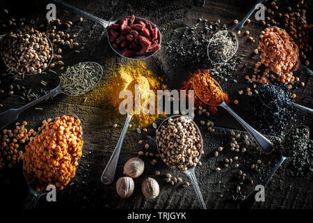 Selezione di spezie su antichi cucchiai d'argento, curcuma, lenticchie, semi di senape, noce moscata, cumino Semi di papavero contro un legno scuro colpo di sfondo da sopra Foto Stock