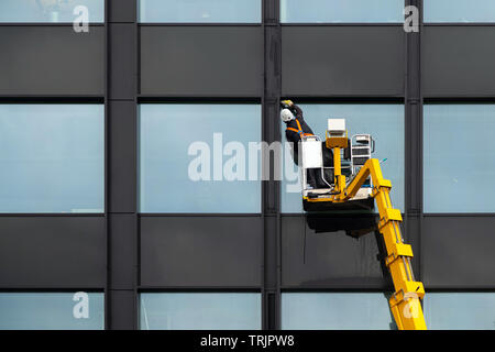 Maschio di detergente per vetri Pulizia vetro windows su edificio moderno in aria in alto su una piattaforma di sollevamento. Lavoratore di vetro di lucidatura in aria in alto Foto Stock