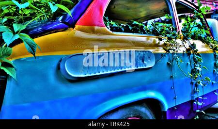 Un vecchio italiano Fiat 500 vettura dipinta nei colori dell'arcobaleno, sommerse in piante rampicanti, parcheggiato su un marciapiede. Foto Stock