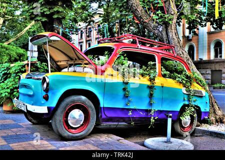 Un vecchio italiano Fiat 500 vettura dipinta nei colori dell'arcobaleno, sommerse in piante rampicanti, parcheggiato su un marciapiede. Foto Stock