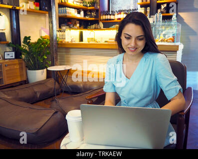 Bellissima gara di misto donna seduta in una caffetteria e sorridenti mentre si utilizza il suo computer portatile Foto Stock