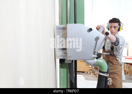 Carpenter uomo lavorare in falegnameria, il taglio di un pannello in legno con circolare verticale macchina sezionatrice, protetto con cuffie e occhiali di protezione Foto Stock