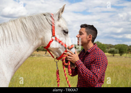 Vista laterale del giovane maschio in camicia a scacchi toccando delicatamente la testa del cavallo bianco con red briglia permanente, mentre in campagna su campo giorno nuvoloso Foto Stock