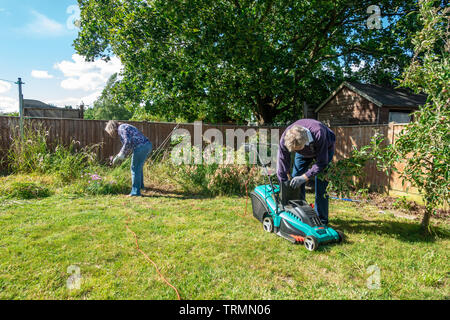 Una coppia in pensione facendo qualche giardinaggio in un giardino residenziale. La signora esegue alcune operazioni di diserbatura mentre l'uomo mows il prato con un tosaerba clectric. Foto Stock