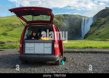 Giovane rilassante in un minivan rosso in sacchi a pelo con apertura dello sportello posteriore. Skogafoss cascata in background, Islanda Foto Stock