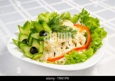 Insalata di pesce - calamari, bastoncini di granchio, cetrioli, uova, la maionese, formaggio, lattuga, pomodoro e olive verdi sul piatto bianco. Foto Stock