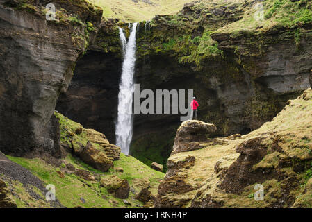 Cascata Kvernufoss nella gola, Islanda. L'uomo turistico in giacca rossa esamina il flusso di caduta in acqua Foto Stock