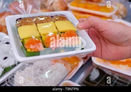 Thai dessert tradizionale, la mano che regge il dolce di riso appiccicoso e rabboccato con uova al vapore con crema pasticcera, pesci secchi e gamberi fritti in scatola di plastica. Foto Stock