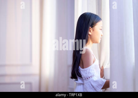 9 anno-piccola vecchia ragazza asiatica in piedi di fronte alla finestra a guardare fuori Foto Stock