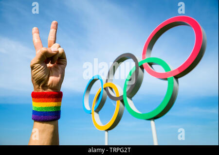 Londra - 4 Maggio 2019: Una mano che indossa il Gay Pride arcobaleno braccialetto colorato fa un segno di pace di fronte anelli olimpici in piedi sotto il luminoso cielo blu. Foto Stock