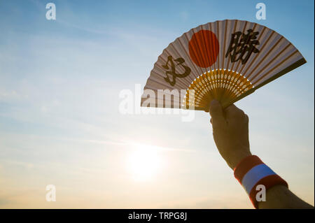Mano del giapponese sostenitore dello sport tenendo una ventola decorate con i caratteri kanji compitazione hissho (traduzione inglese: vittoria certa) di sunrise Foto Stock