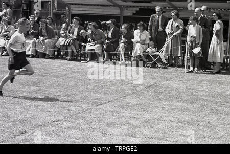 1955, storico, un giovane ragazzo in una scuola primaria la giornata dello sport che sta per attraversare il nastro di rifinitura prima in uno sprint gara di corsa, guardato dai genitori dei bambini, Inghilterra, Regno Unito. Foto Stock