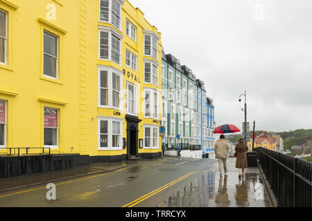 Un paio di camminare lungo la passeggiata di Tenby passato gli edifici colorati con un ombrello in un giorno di pioggia, Pembrokeshire, Galles Foto Stock