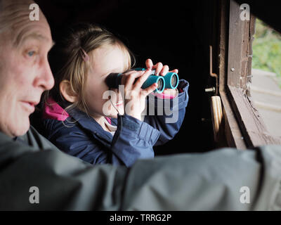 Il Toddler girl utilizza binocolo con uomo più anziano, nonno, in bird-watching nascondere - Carlton, Barnsley, Regno Unito Foto Stock