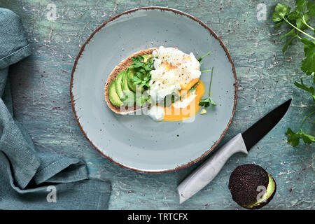 Avocado sandwich con pane scuro, tagliata a fette di avocado e uovo in camicia, piatto giaceva sul turchese sfondo rustico Foto Stock