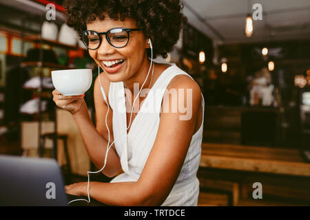 Giovane donna africana a bere caffè e utilizzando il portatile in un cafe'. Donna sorridente seduti al bar a bere caffè e guardare sul computer portatile sul tavolo. Foto Stock