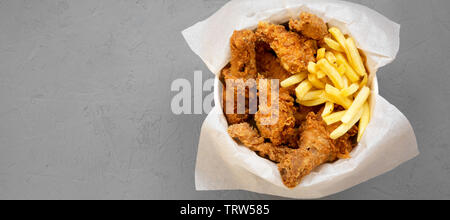 Fritto di pollo Cosce di pollo disossate, piccante ali, patatine fritte e le strisce di pollo in una scatola di carta su uno sfondo grigio, vista dall'alto. Piatto, laici dal di sopra, overhead. Foto Stock
