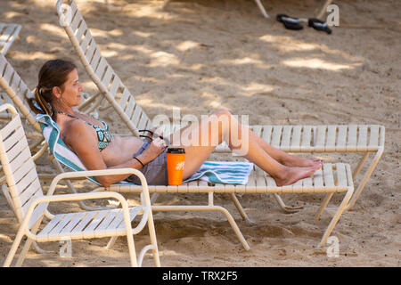 Una donna attraente nel suo 30s, inizio 40s, posa su una sdraio sulla spiaggia a leggere un libro. Foto Stock