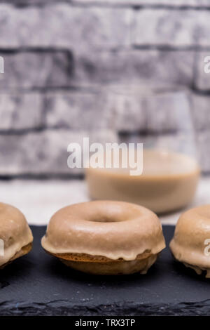 Golden Ginger ciambelle immerso nella Irish Cream smalto con un bicchiere di Irish Cream dietro e una superficie strutturata, grigio Sfondo mattone Foto Stock