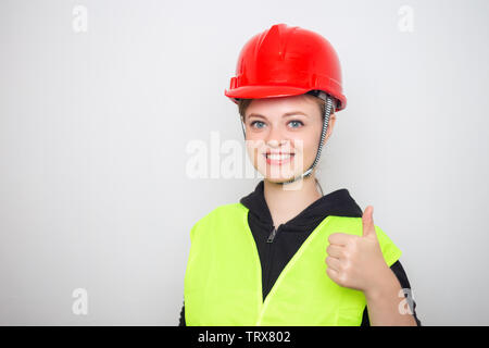 Giovane donna caucasica indossando sicurezza rosso di elmetto e giubbotto, sorridente con il pollice in alto Foto Stock