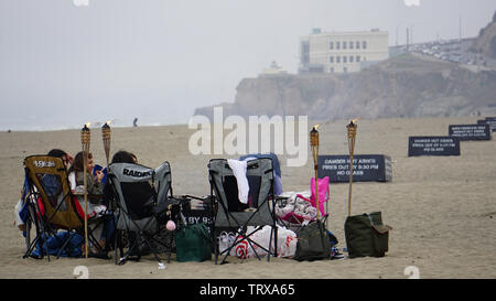 Amici riuniti sulla spiaggia dell'oceano con sdraio e lettini e torce tiki su un nebbioso giorno. Cliff House in background. San Francisco, California, Stati Uniti d'America Foto Stock