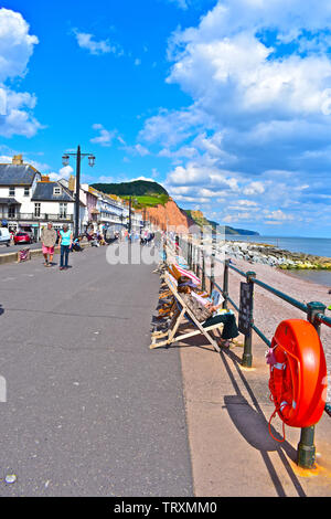 Le persone sedute a sdraio lungo la passeggiata sul lungomare di Sidmouth.Gli altri godere di passeggiate lungo il fronte mare con Jurassic costa dietro Foto Stock