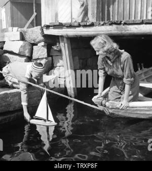 1940s'estate. Una giovane donna e un ragazzo su un giorno d'estate. Il ragazzo sta giocando con un modello di barca a vela e lo inserisce nell'acqua. La Svezia 1946 Kristoffersson ref AC136-5. Foto Stock