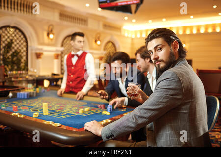 Un giovane uomo bruna in un vestito con un bicchiere di whiskey in mano seduto al tavolo della roulette giocando a poker in un casinò. Foto Stock