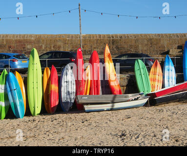 Tavole da surf, canoe e kayak che si asciugano sulla spiaggia al sole del pomeriggio a Mousehole, Cornovaglia, Inghilterra, Regno Unito sotto un cielo blu. Foto Stock