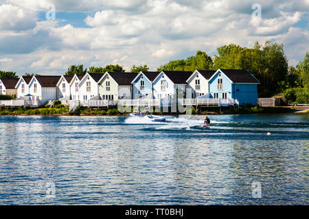 Un sciatore acqua scorre oltre la Luxury Holiday Homes intorno al Lago di primavera nel Cotswold Water Park, Sud Cerney, nel Gloucestershire. Foto Stock