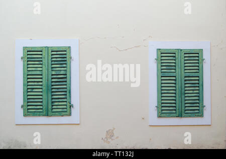 Due vecchie finestre vengono chiuse con persiane verdi contro il muro beige. Foto Stock