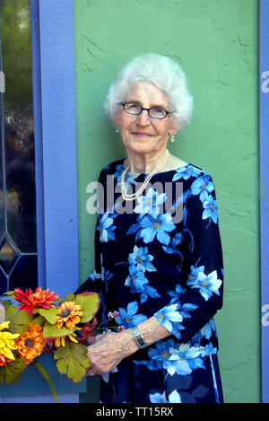 Elegante donna anziana organizza fiori in una scatola di vetro al di fuori del suo negozio di articoli da regalo. Indossa un abito e perle e un lieve sorriso. Foto Stock