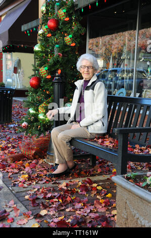 Piuttosto la nonna si siede su una panca in legno appoggiata da shopping di Natale. Albero di natale con le luci si siede accanto alla sua downtown banco. Foto Stock