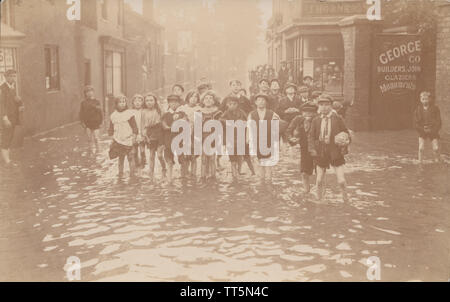 Vintage Edwardian Cartolina fotografica che mostra un folto gruppo di bambini che giocano nelle acque di esondazione in una cittadina britannica o strada di città, Foto Stock