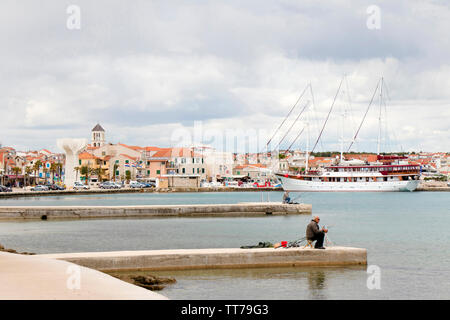 Vodice, Croazia - 30 Aprile 2019: due uomini seduti da soli e paziente sul molo sul mare pesca con canne da pesca e una nave a vela ormeggiata dietro di loro w Foto Stock