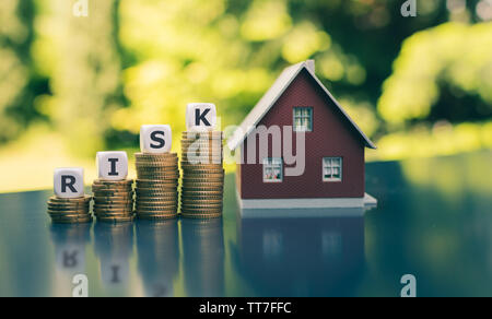 Concetto di rischio finanziario per comprare una casa. Dadi forma la parola "rischio" posto sulla crescente alte pile di monete accanto a un modello di casa. Foto Stock
