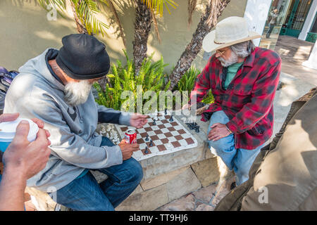 Un vecchio maschio caucasico detiene un segno sfidando la gente a un gioco di scacchi. Egli ha la sua scacchiera impostare pronto per la riproduzione su un basso muro di pietra. Foto Stock