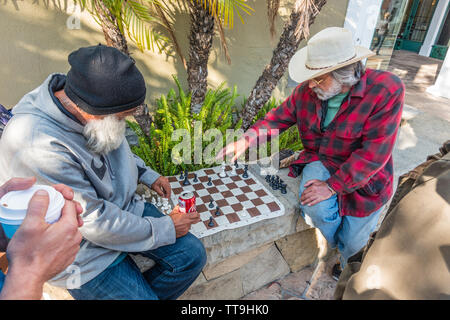 Un vecchio maschio caucasico detiene un segno sfidando la gente a un gioco di scacchi. Egli ha la sua scacchiera impostare pronto per la riproduzione su un basso muro di pietra. Foto Stock