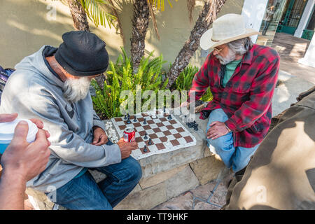 Un vecchio maschio caucasico detiene un segno sfidando la gente a un gioco di scacchi. Egli ha la sua scacchiera impostare pronto per la riproduzione su un basso muro di pietra. Foto Stock