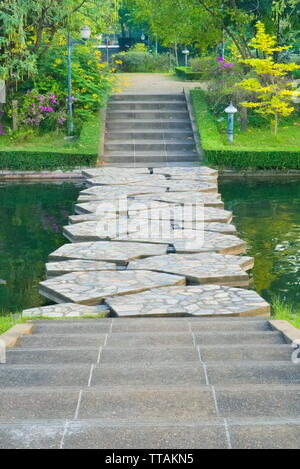 Una vista verticale di cemento gradini che conducono in basso verso un canale verde ponte in pietra con spazi acquosi, e oltre in un delizioso giardino fiorito park. Foto Stock