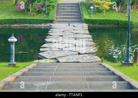 Una vista del paesaggio di cemento gradini che conducono in basso verso un canale verde ponte in pietra con spazi acquosi, e oltre in un delizioso giardino fiorito park. Foto Stock